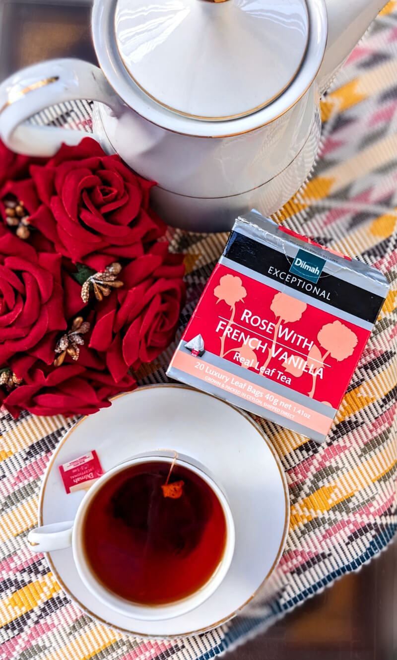 Dilmah's Rose With French Vanilla Tea box next to a teapot, a cup of brewed tea and roses